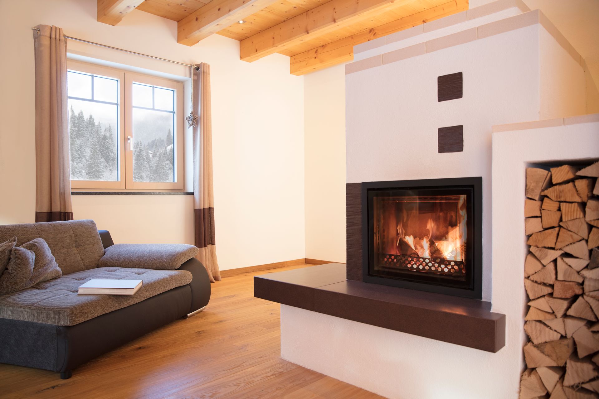 Gemütliches Wohnzimmer mit Sofa, Fenster und einem brennenden Kamin neben gestapeltem Brennholz.