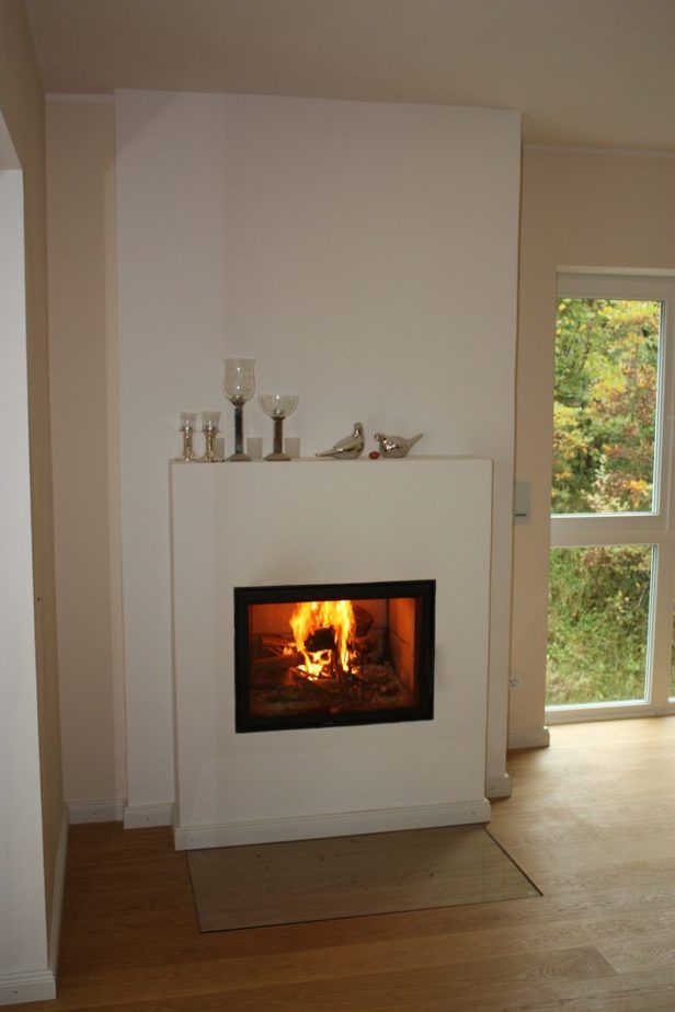 Wohnzimmer mit weißem Kamin und brennendem Feuer, neben einem hohen Fenster mit Blick ins Grüne.