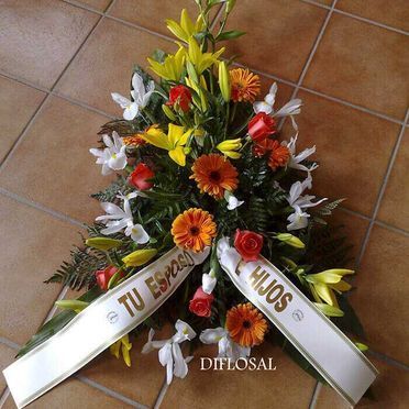 Arreglo floral con flores naranjas, amarillas y blancas, con cintas que dicen