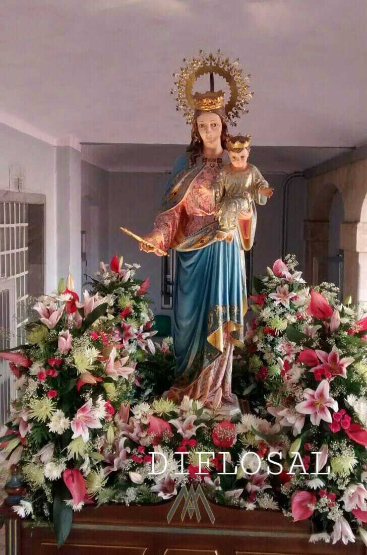 Estatua de la Virgen María sosteniendo a un niño, rodeada de arreglos florales, en un edificio.