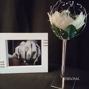 Foto enmarcada en blanco junto a una flor decorativa en un soporte. La foto muestra las manos unidas; la flor tiene pétalos blancos.