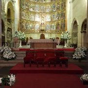 Interior de una catedral con un altar, un fondo dorado ornamentado, alfombra roja y arreglos florales blancos.