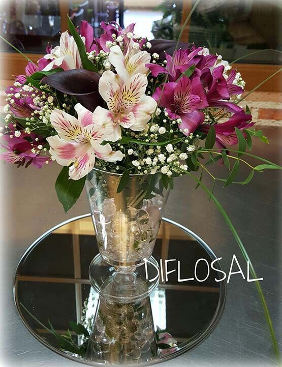 Arreglo floral en jarrón de vidrio con flores blancas y moradas, apoyado sobre una base de espejo.