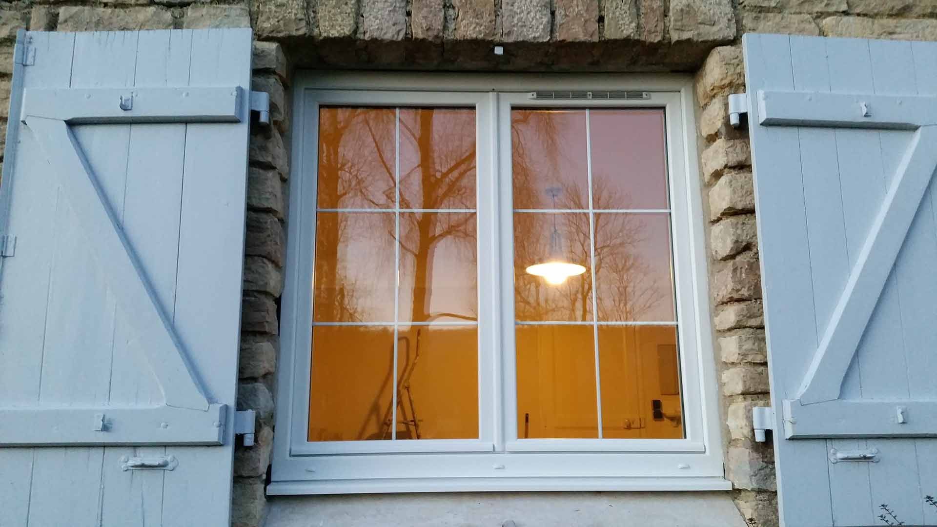 Fenêtre à volets blancs, illuminée d'une chaude lueur jaune dans un mur de pierre au crépuscule