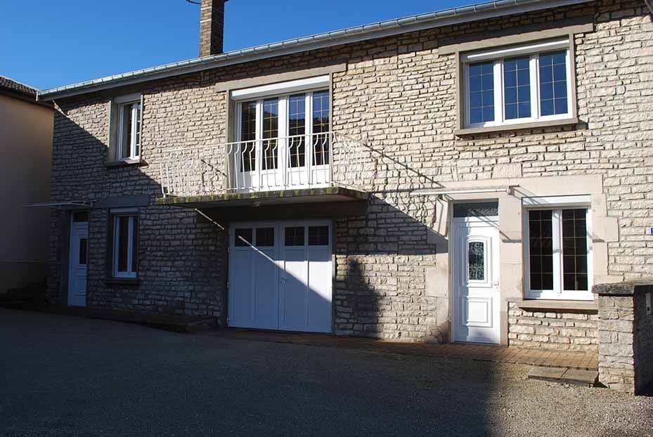 Maison en pierre avec balcon, porte de garage et fenêtres à cadre blanc baignées de soleil