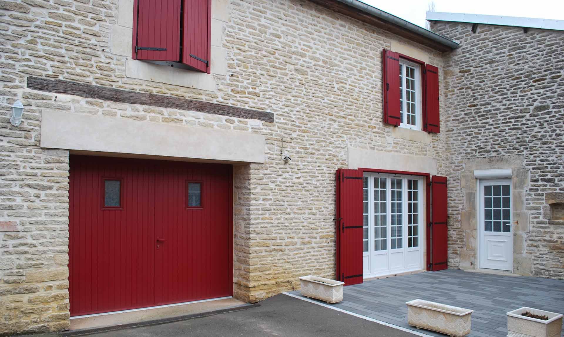Façade en pierre d'une maison avec garage et volets rouges, fenêtres à encadrement blanc et cour pavée.