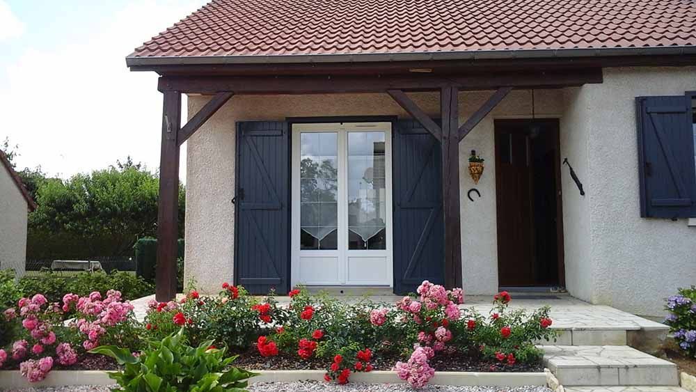 Petite véranda avec volets bleus, double porte blanche et jardin fleuri devant.
