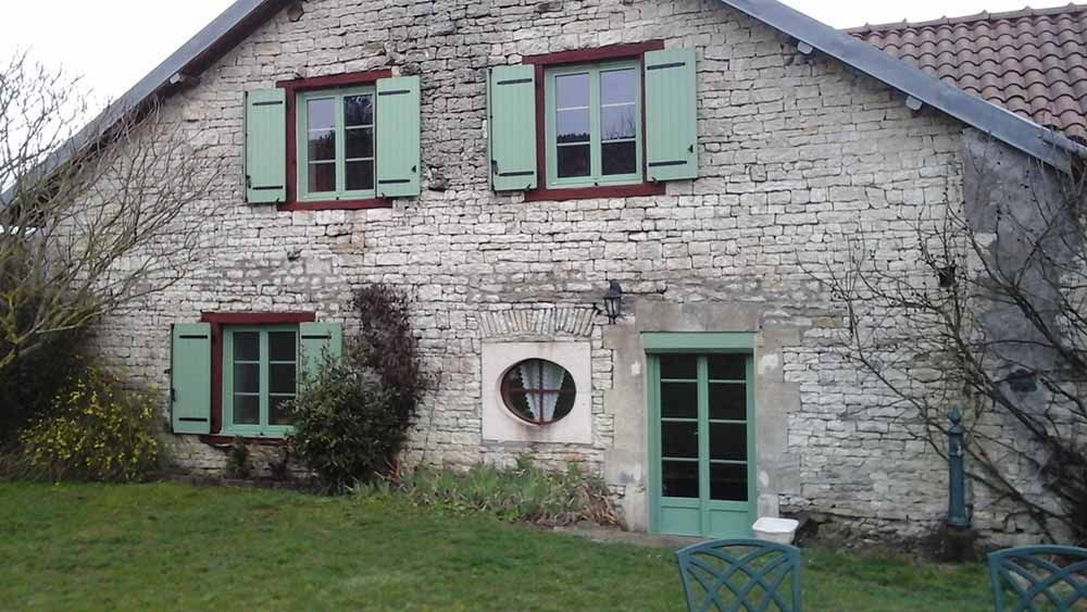 Maisonnette en pierre blanche avec volets et porte verts, pelouse devant et fenêtre ronde au-dessus de l'entrée.