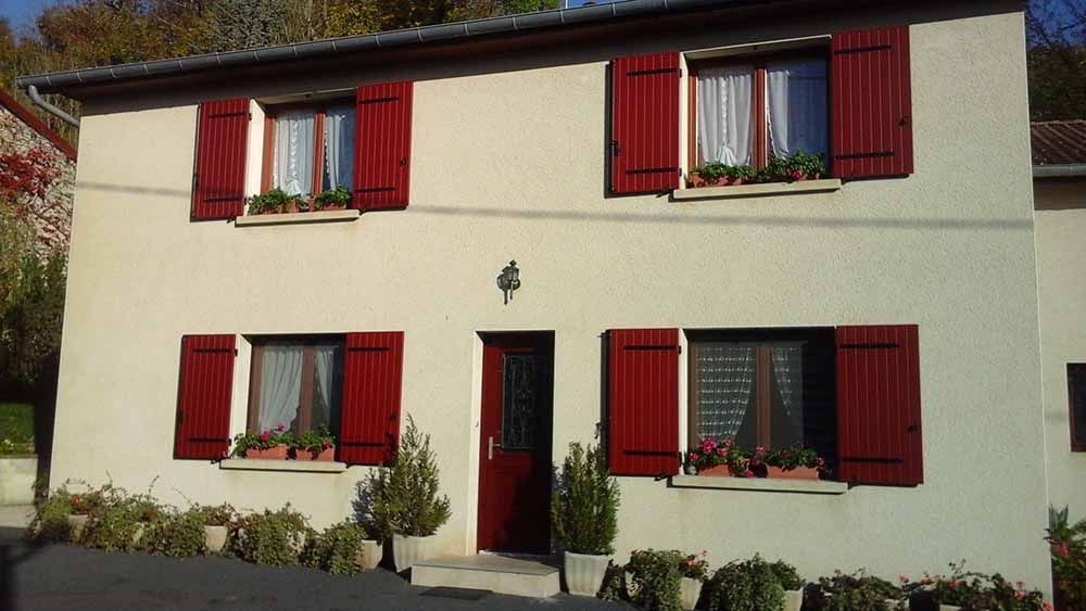 Maison crème à deux étages avec volets rouges et porte d'entrée rouge, entourée de plantes de jardin.