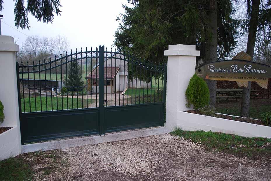 Un portail en fer forgé vert marque l'entrée d'un bâtiment fermé par un portail, avec des piliers blancs et un panneau en bois sur la droite.