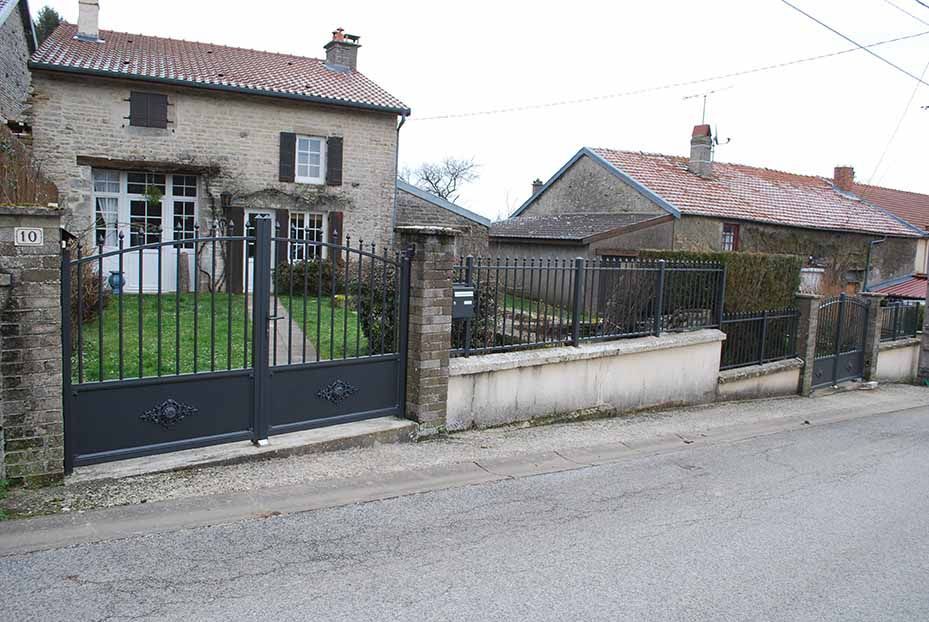 Maison en pierre avec portail en fer noir et muret bas, située dans une rue résidentielle calme.