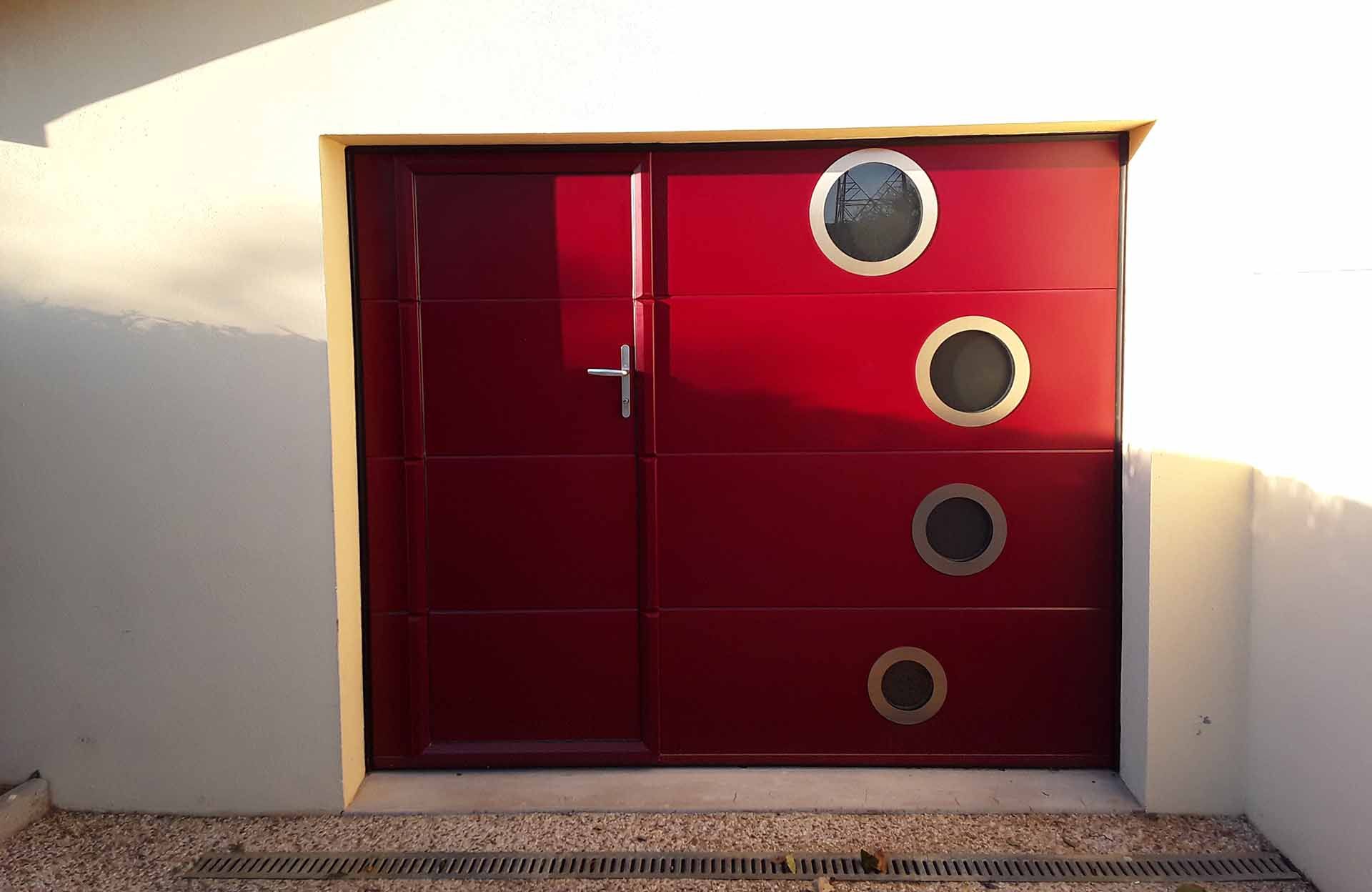 Double porte rouge avec fenêtres circulaires dans un mur blanc, éclairée par la lumière du soleil.