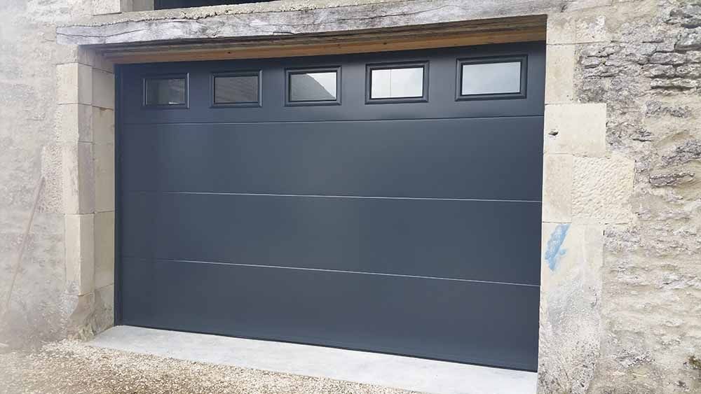 Porte de garage grise encastrée dans un mur en pierre, avec quatre petites fenêtres en partie supérieure.