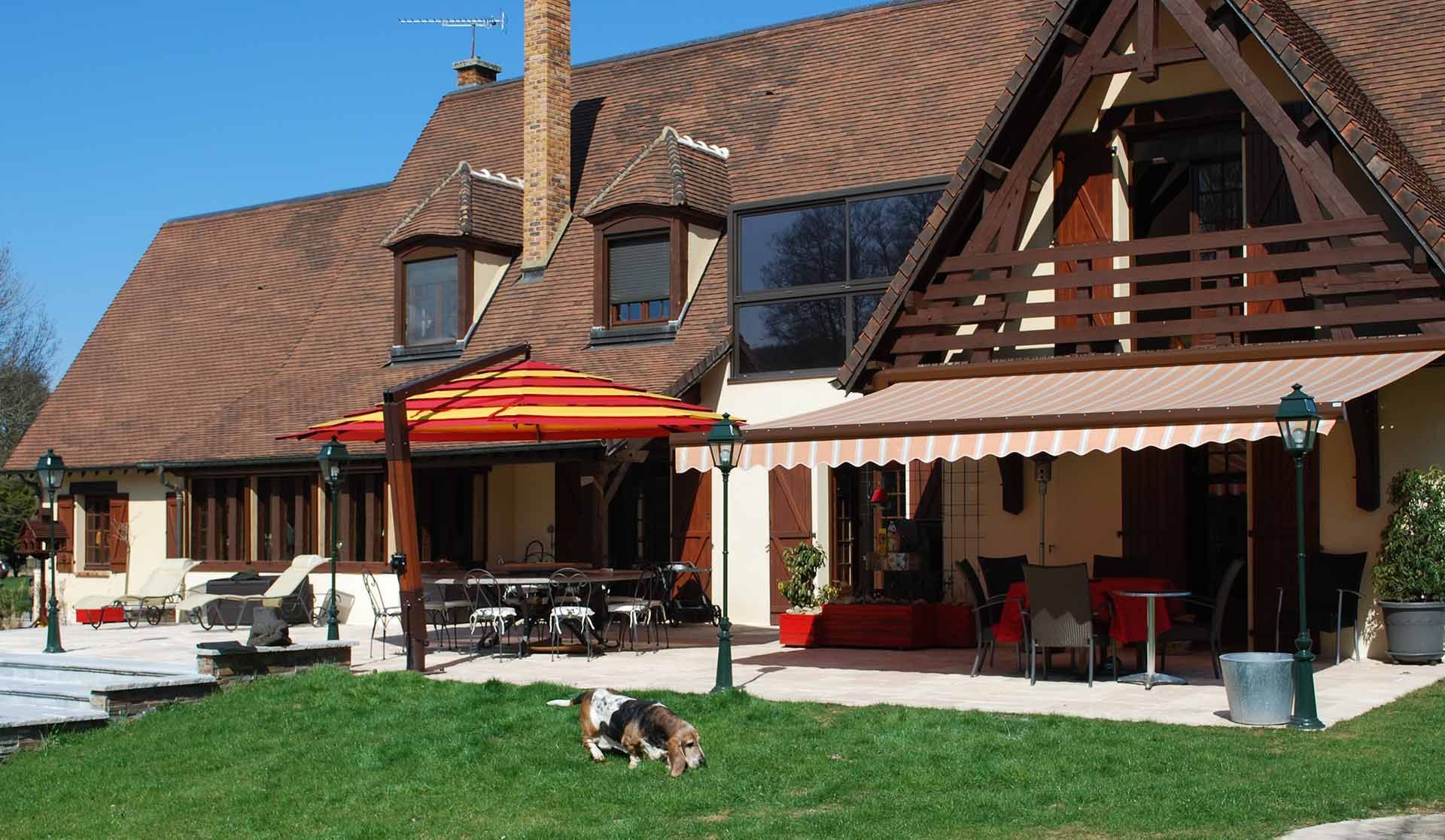 Grande maison au toit de tuiles rouges, parasols de terrasse et petit chien sur une pelouse verte