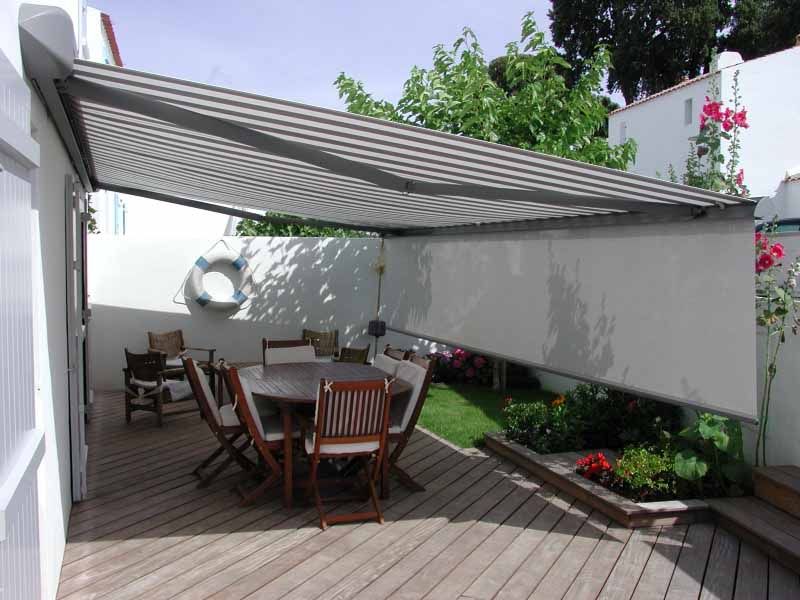 Terrasse extérieure ombragée avec table et chaises en bois, et auvent rétractable, jouxtant un mur blanc et un parterre de fleurs.