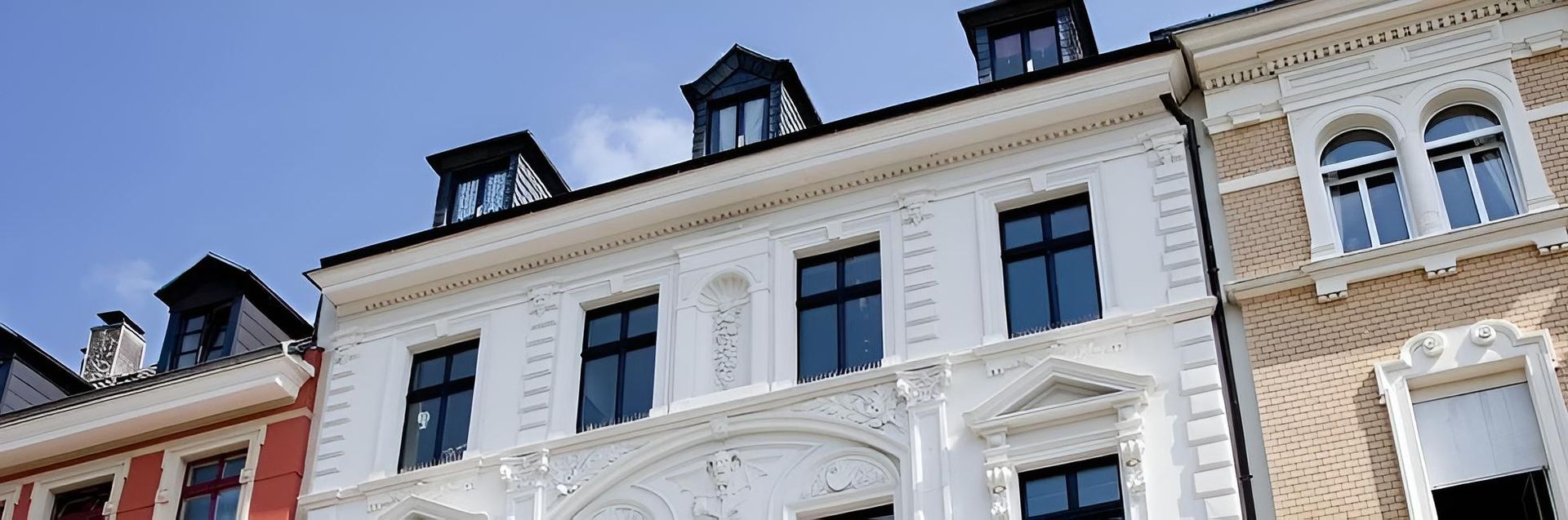 Weisse historische Gebäudefassade mit Bogenfenstern und schwarzen Dachgauben vor blauem Himmel