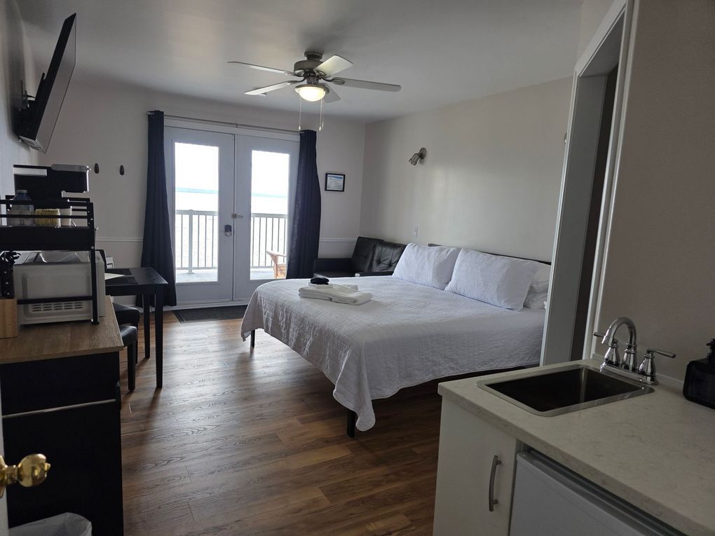 Bedroom with a neatly made white bed, wood floor, TV, and balcony doors with curtains
