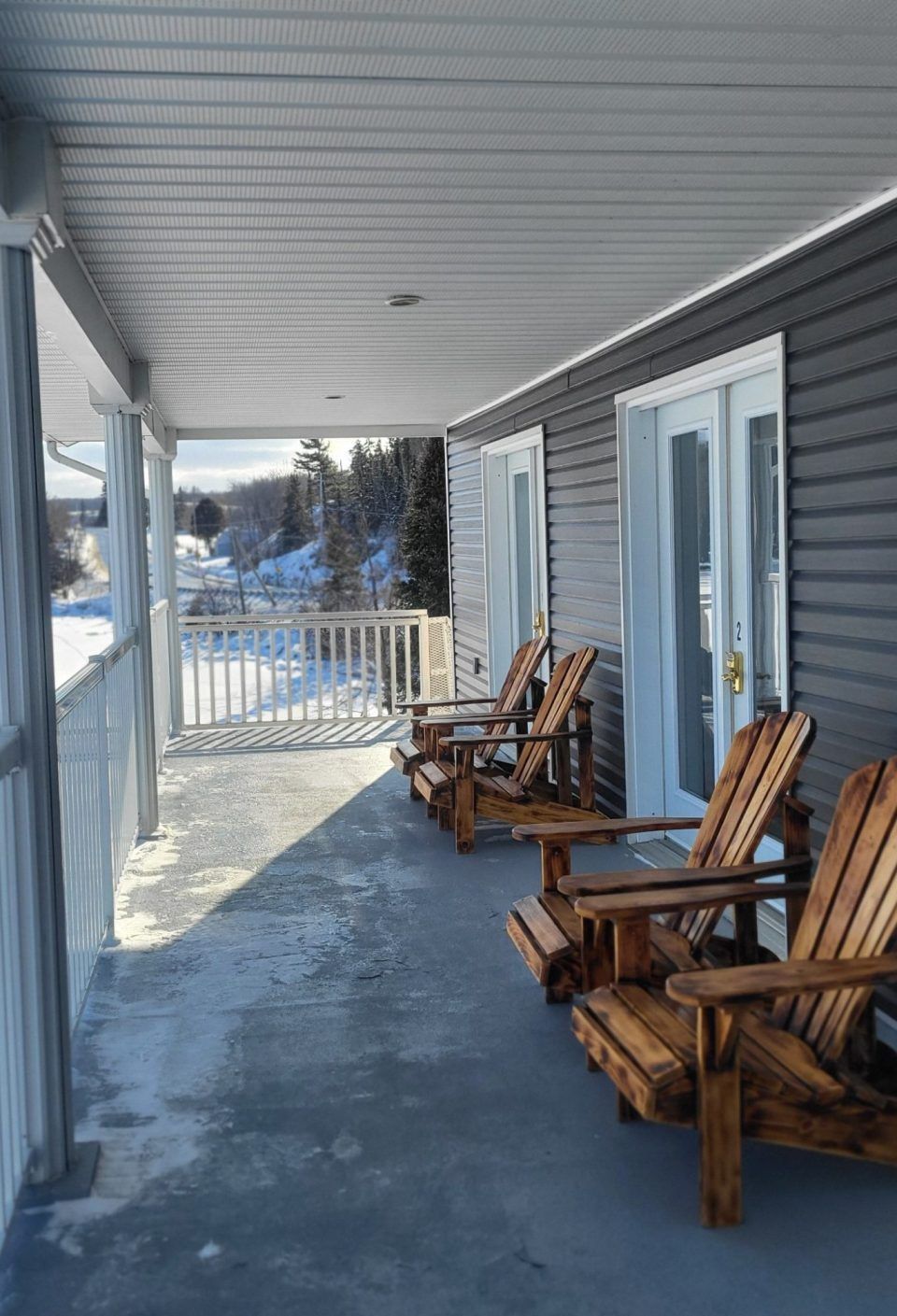 Snowy porch with wooden chairs and white railings beside a gray house