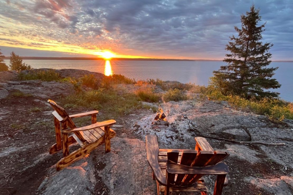 Rocky lakeshore at sunset with two wooden chairs around a fire pit overlooking calm water