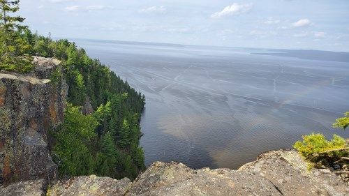 Cliffside overlook above a wide gray lake or river, with green trees and rocky foreground under a hazy sky