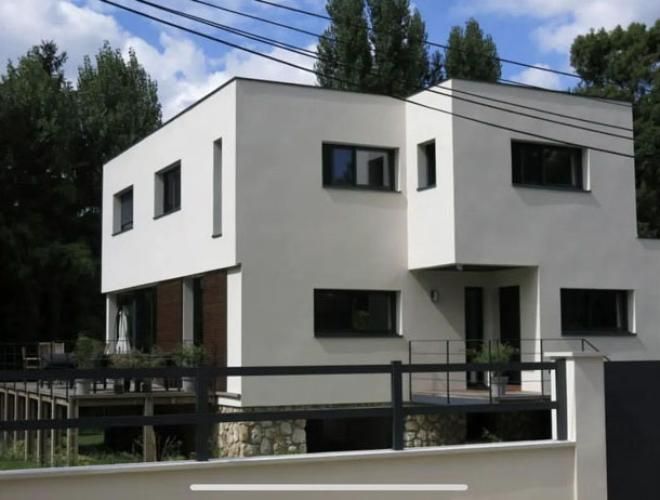 Maison moderne blanche à deux étages, avec des menuiseries foncées, des balcons et des arbres en arrière-plan