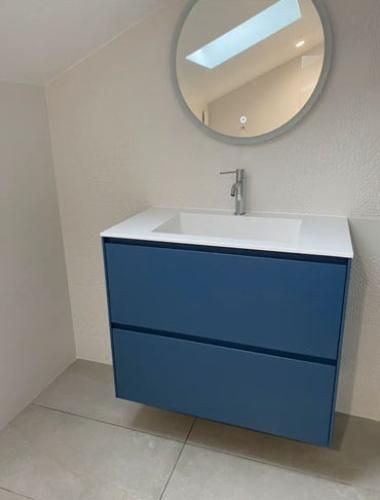 Meuble-lavabo moderne avec des tiroirs bleus, un lavabo blanc, un miroir rond et un sol en carrelage clair.
