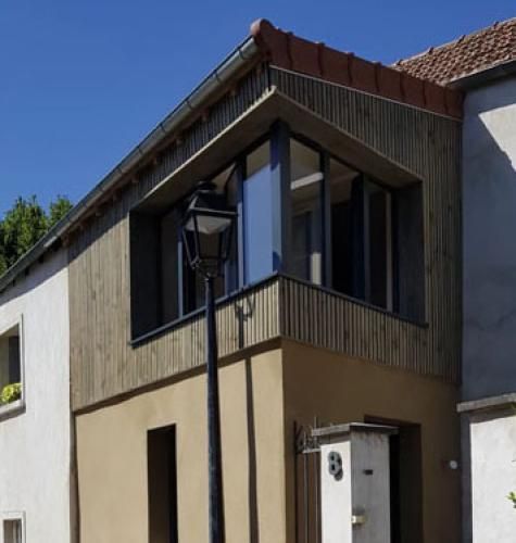 Maison à deux étages avec des murs beiges, des fenêtres sombres sur le balcon et un lampadaire devant.