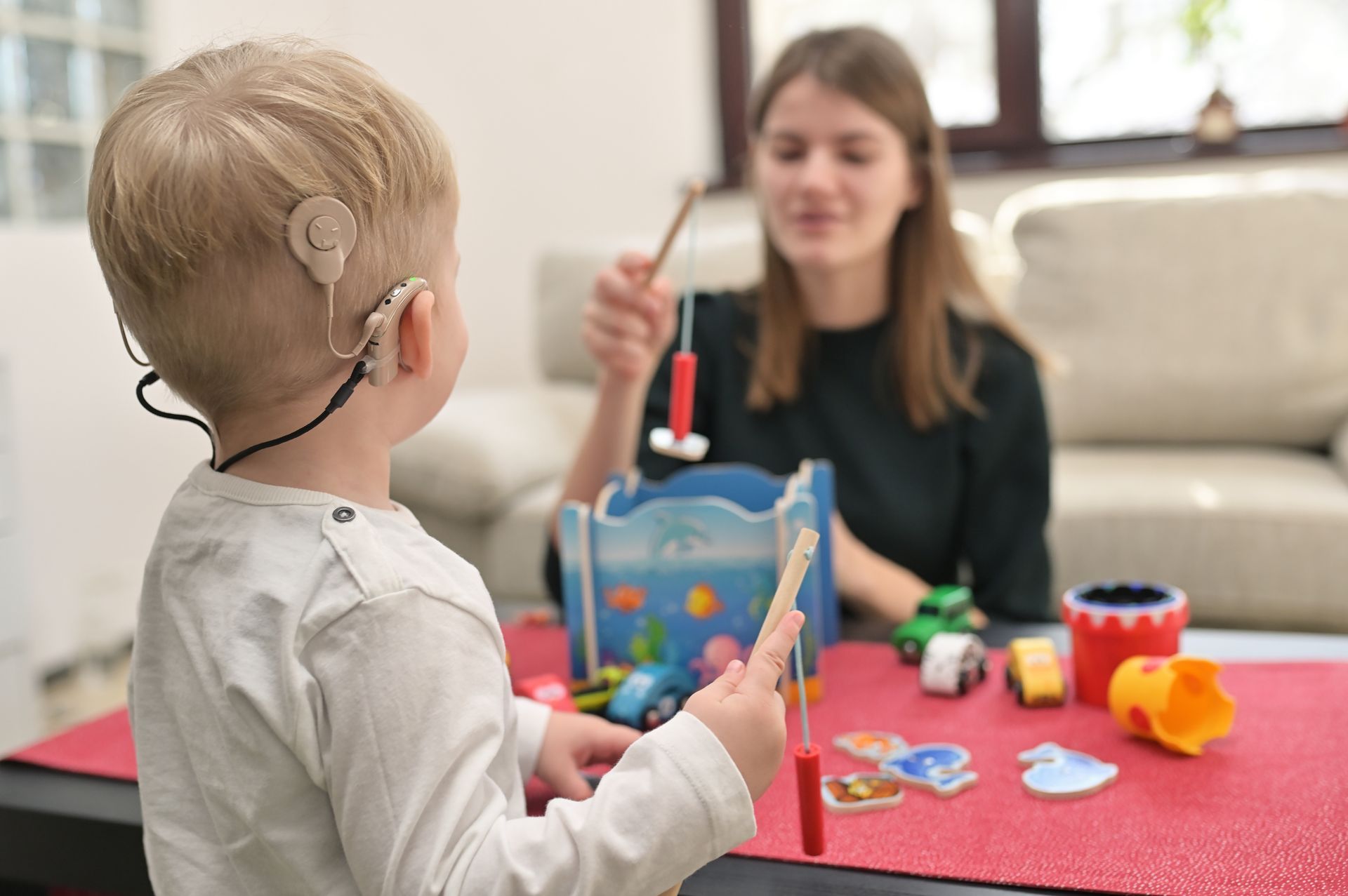 Ein kleiner Junge mit Hörgerät spielt mit Spielzeug, während eine Frau zusieht.