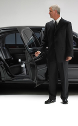 Un chófer con traje negro de pie junto a la puerta abierta de un coche en un escenario blanco iluminado como un estudio.