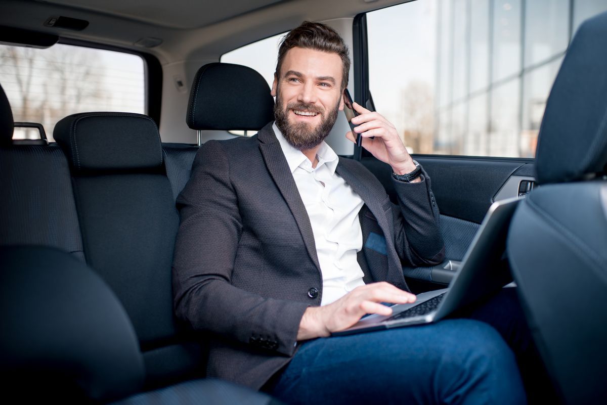 Un profesional sonriente, usa un ordenador portátil mientras habla por teléfono en el asiento trasero de un coche.