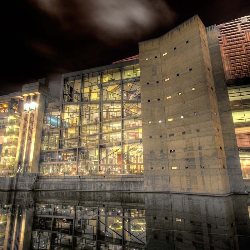 Edificio moderno de vidrio y hormigón iluminado por la noche, reflejándose en aguas tranquilas.
