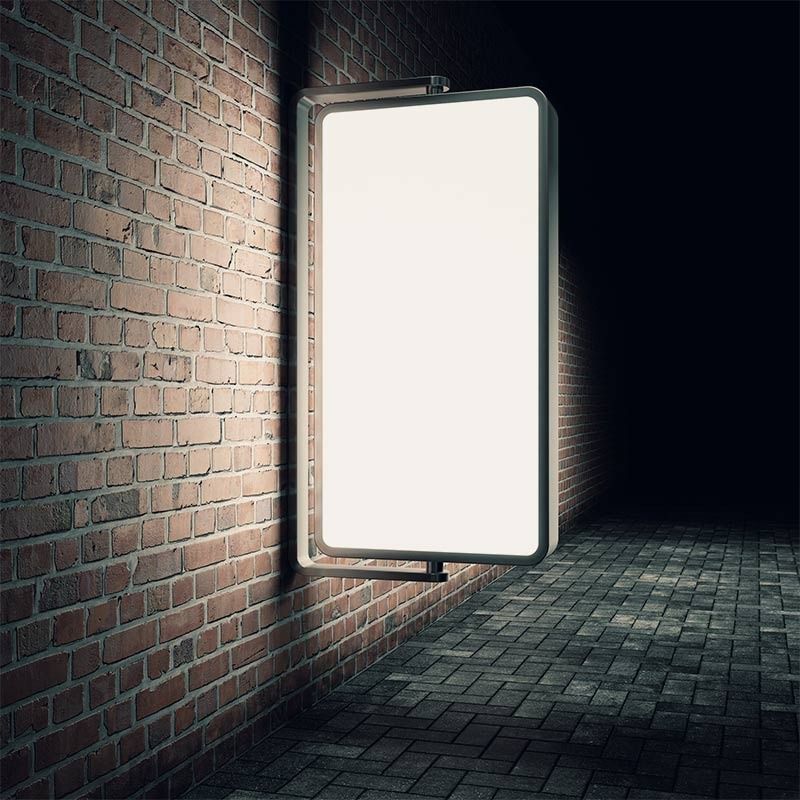 Un cartel publicitario brillante y en blanco montado en una pared de ladrillos por la noche en una calle pavimentada.