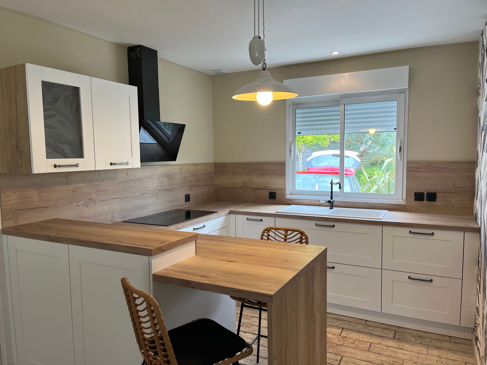 Cuisine blanche et boisée avec placards hauts, îlot central, hotte noire et fenêtre avec vue sur l'extérieur.