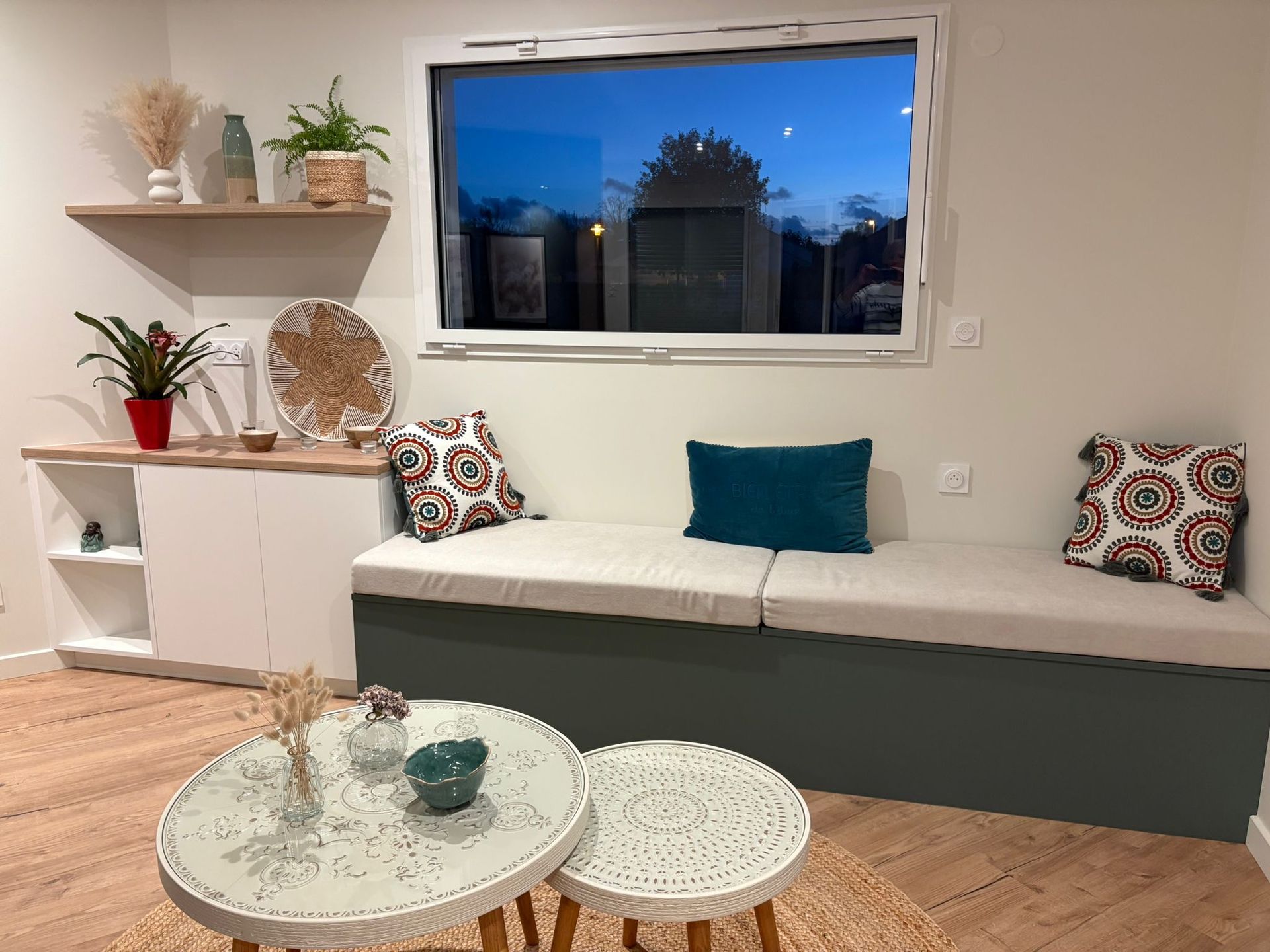 Salon confortable avec banquette intégrée, deux tables basses rondes et une fenêtre donnant sur le ciel du soir.