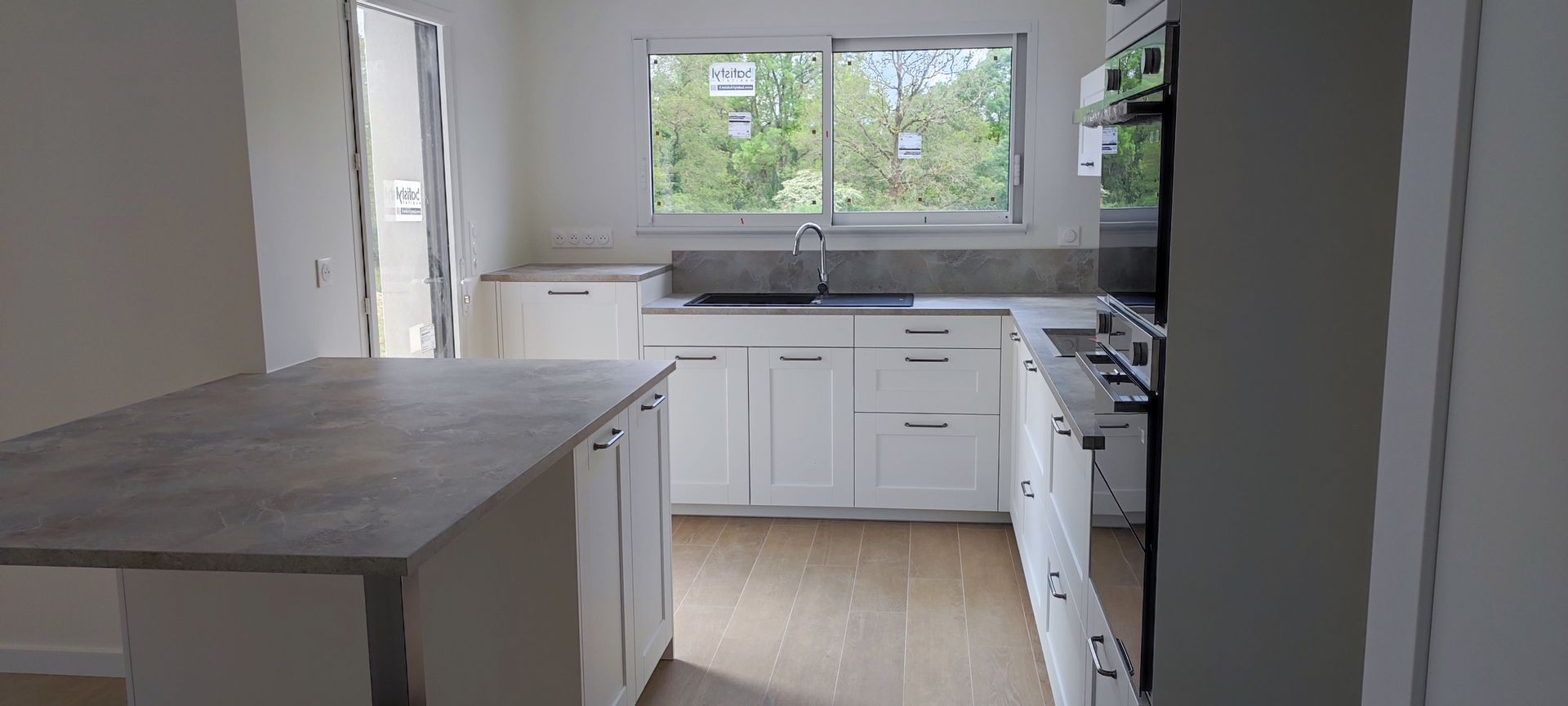 Cuisine moderne avec placards blancs, plan de travail gris et îlot central. Une fenêtre donne sur les arbres à l'extérieur.