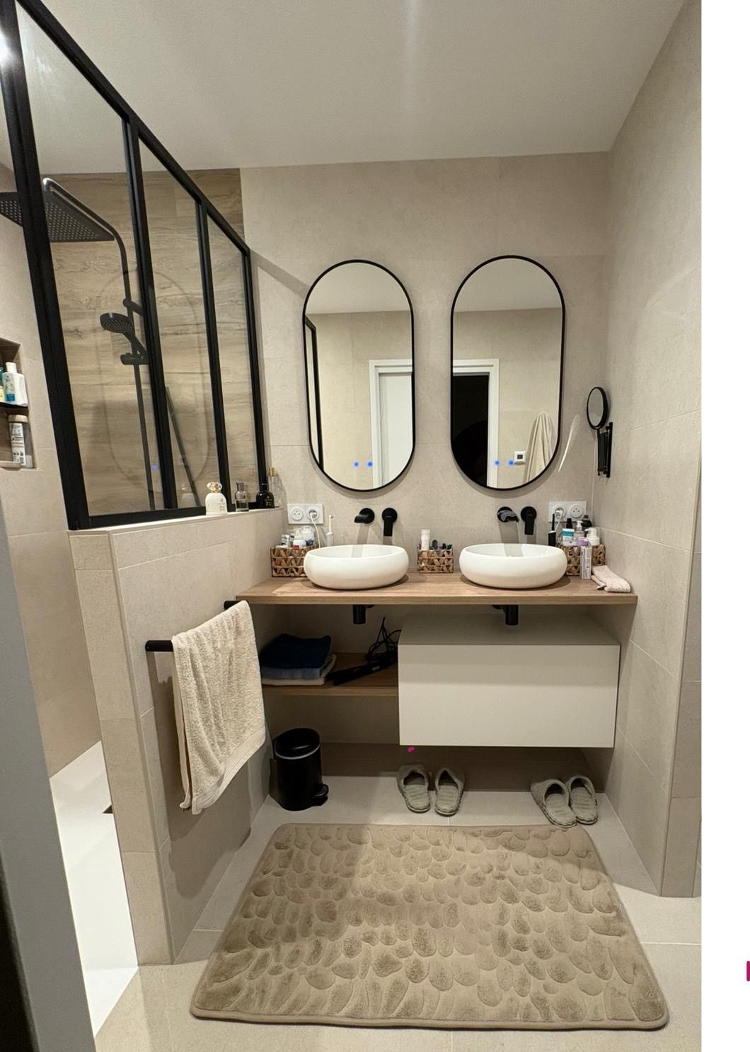 Salle de bains moderne avec deux lavabos, miroirs ovales, espace douche et tons beiges.