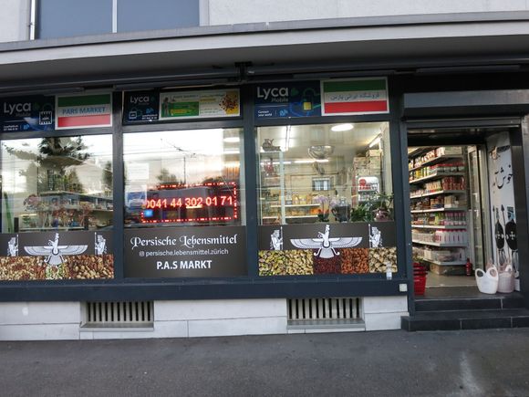 Exterior view of a Persian grocery store with a glass front, displaying Iranian flags and various goods.