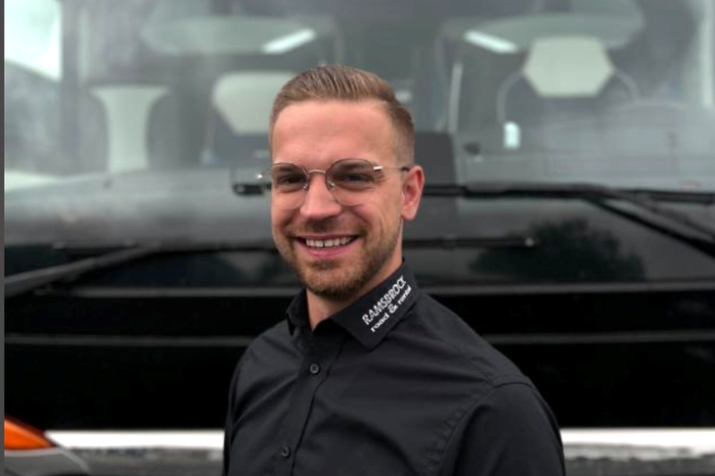 Ein Mann mit Brille und schwarzem Hemd steht lächelnd vor einem Bus.