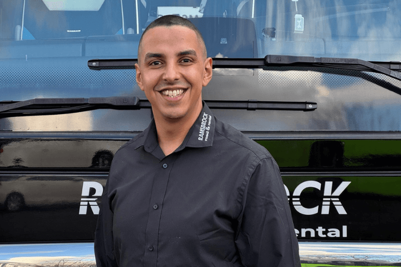 Ein Mann in einem schwarzen Hemd lächelt vor einem Bus.