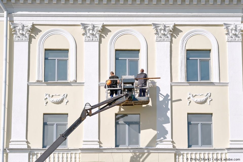 Zwei Arbeiter reparieren auf einer Hubarbeitsbühne die Fassade eines hellen Gebäudes mit Bogenfenstern.