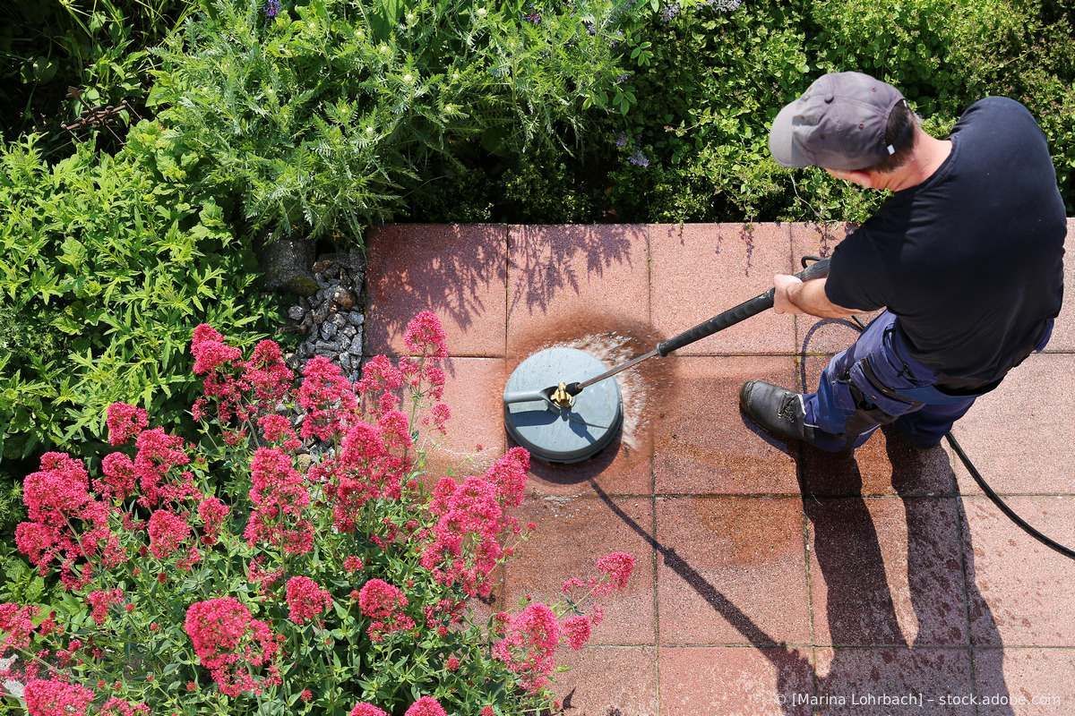 Eine Person reinigt mit einem runden Hochdruckreinigeraufsatz eine rot geflieste Terrasse neben einem blühenden Gartenbusch.