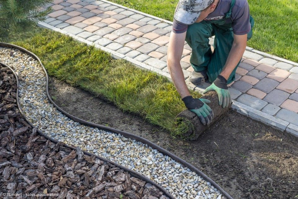 Ein Gärtner kniet auf einem Ziegelweg und legt eine Rasenrolle auf vorbereiteten Boden neben einer Kiesbegrenzung im Garten.
