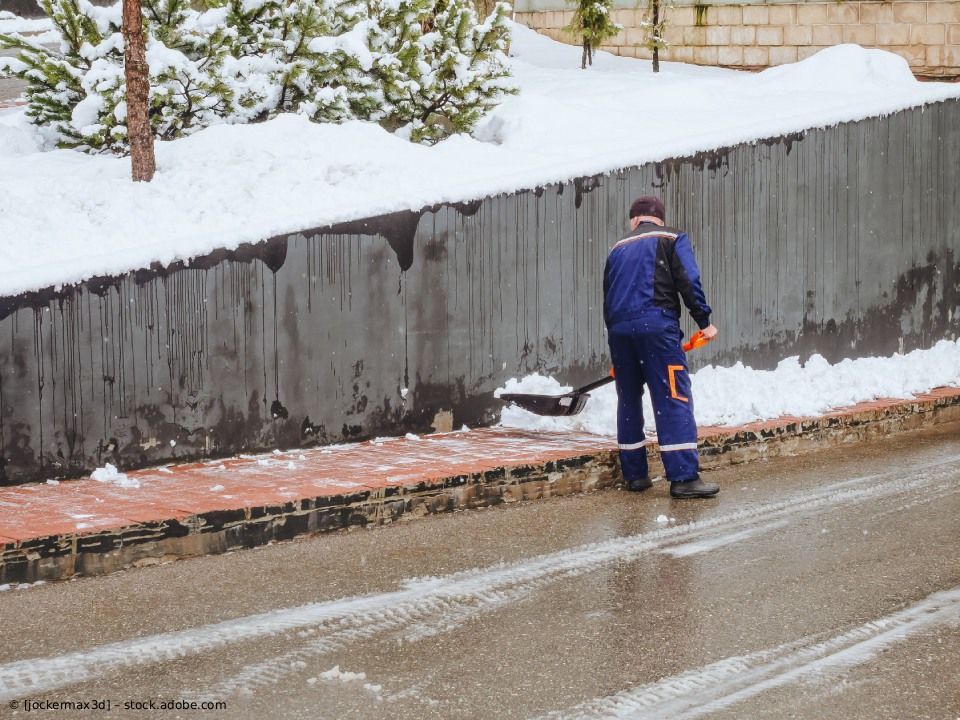 Eine Person in blauer Uniform schaufelt Schnee von einer dunklen Stützmauer auf eine nasse, gepflasterte Straße.