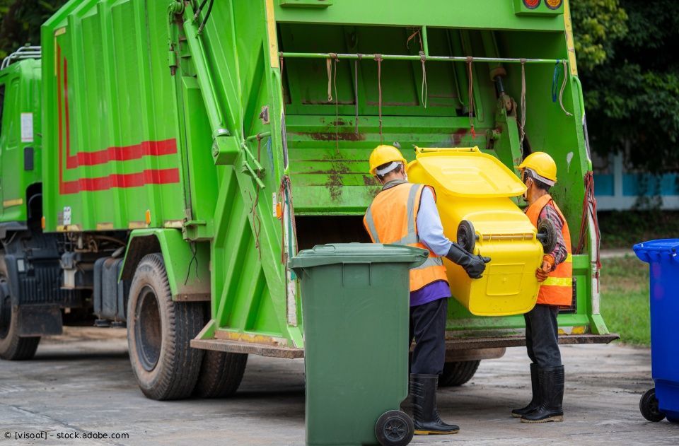Zwei Müllwerker in orangefarbenen Warnwesten und Schutzhelmen verladen einen gelben Müllcontainer in einen grünen Müllwagen.