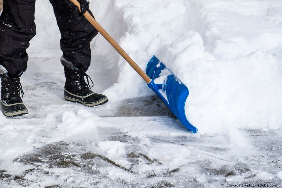 Eine Person, die schwarze Winterstiefel und -hosen trägt, benutzt eine blaue Schneeschaufel,