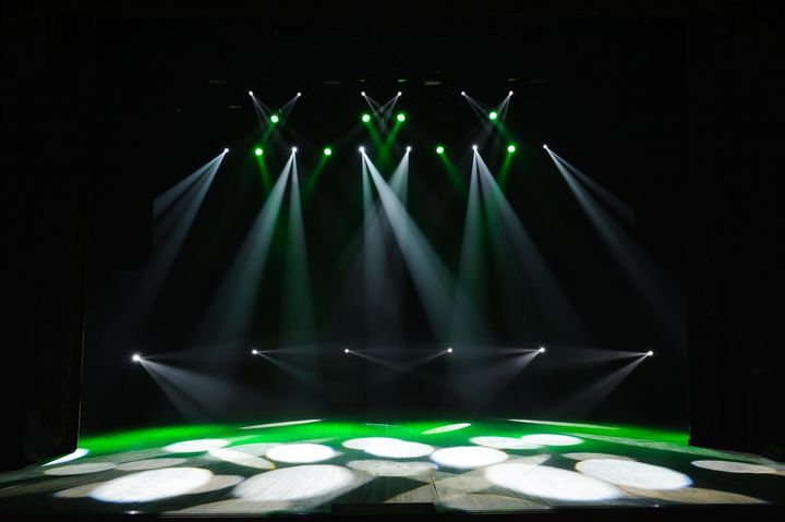 El escenario está iluminado con focos verdes y blancos sobre un fondo negro. El suelo presenta un patrón de círculos blancos.