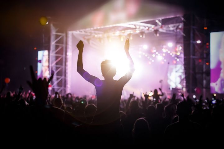 Persona con los brazos en alto entre la multitud en un concierto, recortada contra las brillantes luces del escenario. Otros asistentes al concierto en primer plano.