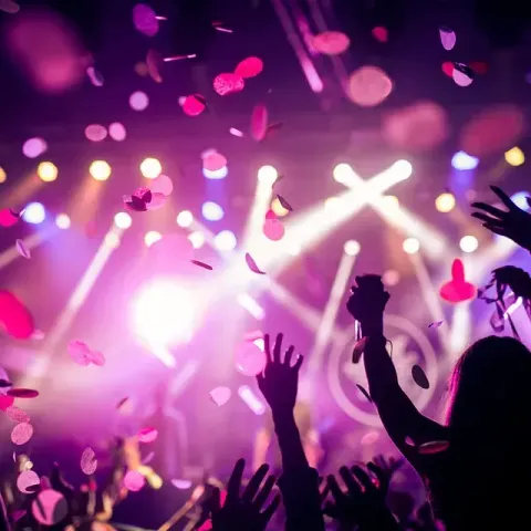 Gente levantando los brazos en un recinto de conciertos abarrotado, con confeti cayendo y luces del escenario brillando en rosa y blanco.