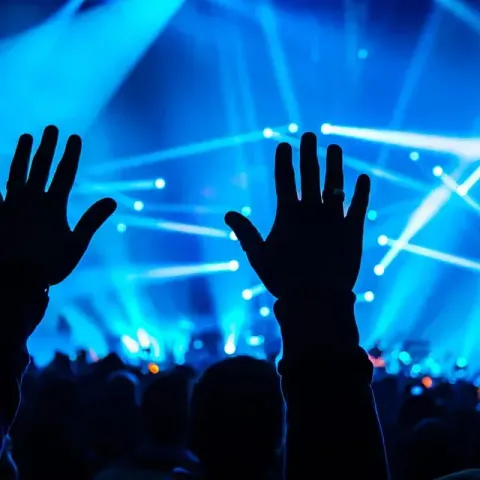 Siluetas de manos alzadas con entusiasmo en un concierto iluminado por focos azules. Multitud al fondo.