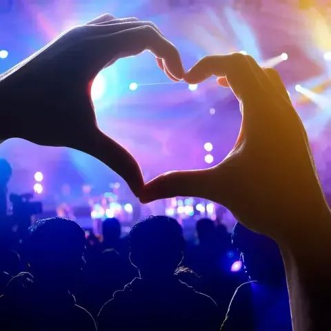 Manos formando un corazón frente a una multitud en un concierto con luces de escenario de colores.